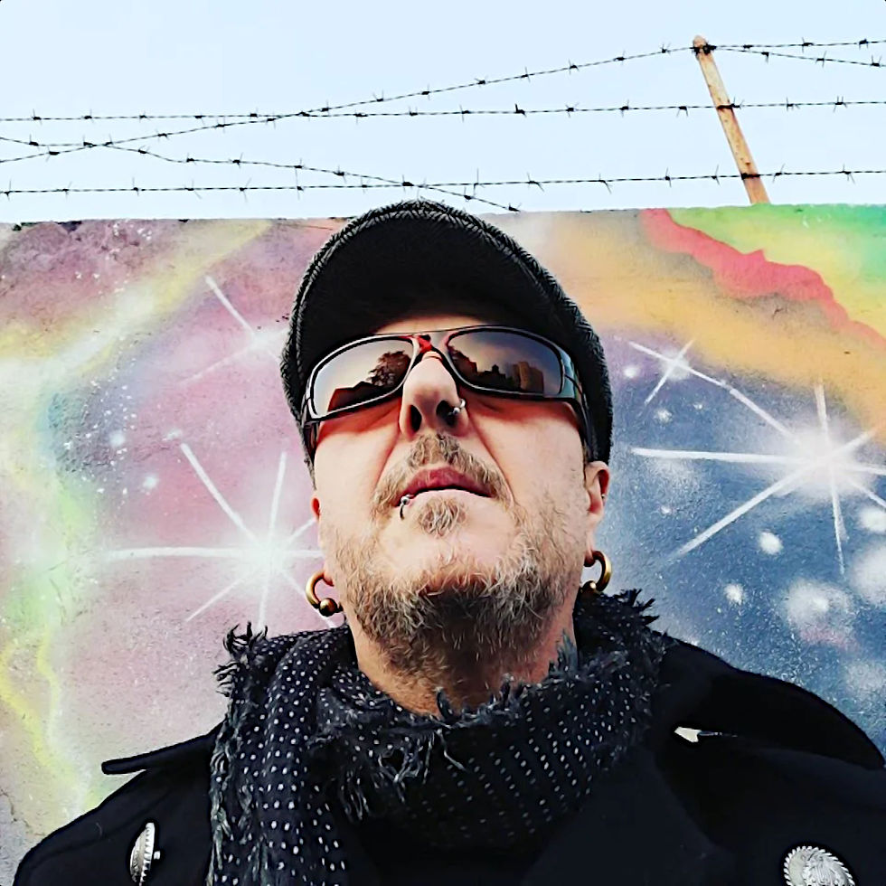 Italian DJ and Producer EL Brujo standing wearing sunglasses in front of graffiti wall topped with barbed wire, colour image.