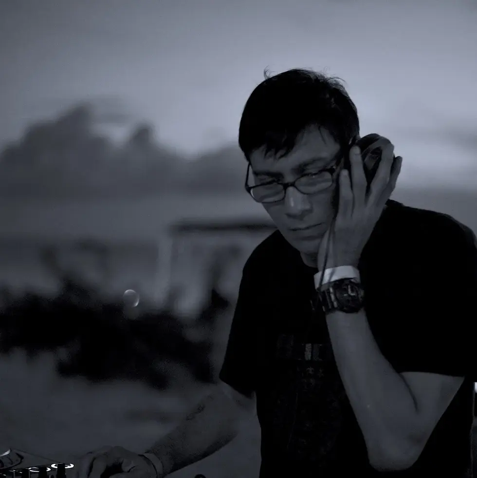 Black-and-white image of Brendon P holding one headphone while performing, representing his long-running work across Singapore’s club scene and international House music circuit.