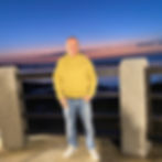 Italian DJ and producer Marco Mei is captured mid-jump with his arms outstretched on the sand dunes of Mui Ne, Phan Thiet, Vietnam. He sports sunglasses, yellow shorts, and a T-shirt in a vibrant, colorful scene.