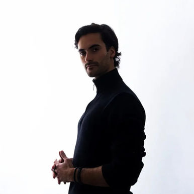 Salvatt standing in a black high-neck top with his hands clasped against a plain white background.