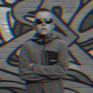Portrait of Kevin Manning, DJ, producer and remixer from Belfast, Northern Ireland, wearing sunglasses and a jacket with arms crossed in front of a graffiti wall with a blur effect.