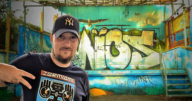French DJ Phoenixx standing in front of a derelict stage with  wall covered in Graffiti in Germany wearing a Yankees baseball cap and holding his fingers in a two fingered salute, colour image