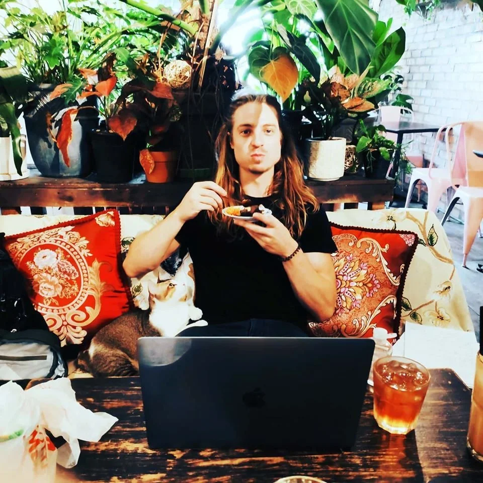British DJ and producer Ezmun sitting in a café with his laptop drinking coffee and eating a cake, colour image.