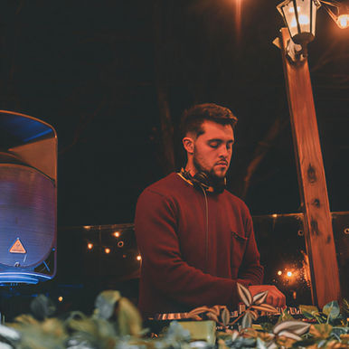 Argentinian DJ and producer Federico Cepeda performing at an outdoor night event, standing behind the decks with headphones around his neck, illuminated by warm lights beside a speaker and wooden lamp post.