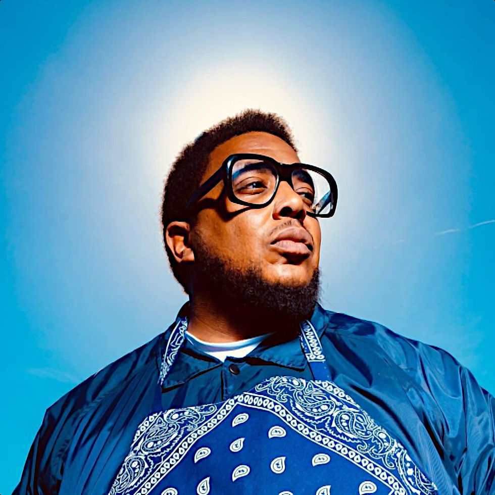 American producer and Rapper Glasses Malone in an outdoor uppper body shot with a blue sky in the background wearing clear big rimmed glasses and looking to his left, colour image.