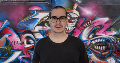 British DJ L-J Louis standing in front of graffiti wall in Melbourne wearing a black T-shirt and glasses, colour image.