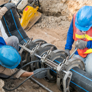 Two workers in blue hard hats and orange safety vests installing a water pipeline at a construction site in Latin America
