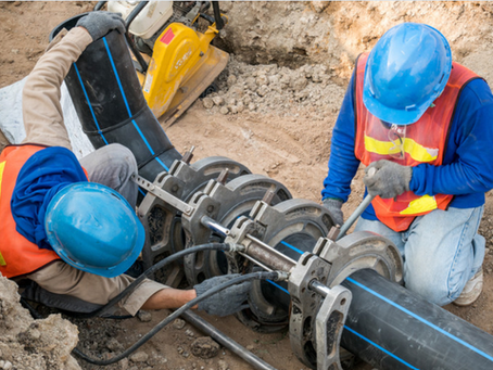 Dos trabajadores con cascos azules y chalecos de seguridad naranjas instalando una tubería de agua en una obra de construcción en América Latina