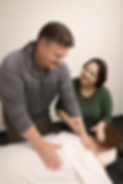 A man is massaging someone who is face down on a massage table. Next to him is a friendly woman instructing him.