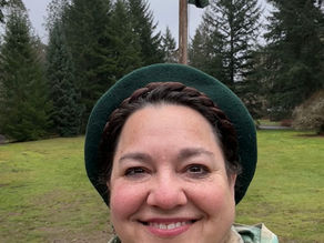 Holostill (photo) of a woman in front of a lush landscape, wearing a camo poncho like Leia on Endor. Her long hair is bound up in Alderaanian braids, and she's wearing a dark green hat.  Behind her is a flagpole with a Selvarin flag. 