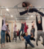 Group of people hanging from metal bars indoors, one woman on top. Casual attire, playful mood, neutral-colored room.