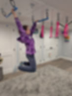 Child in purple hoodie swings on gym rings in a room with white doors. Pink ropes and a small basketball hoop are visible in the background.
