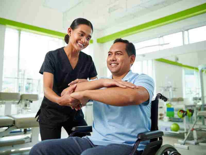 Filipino physical therapist in black scrubs working with a Filipino patient with disability in bright, clean rehabilitation clinic with green accents