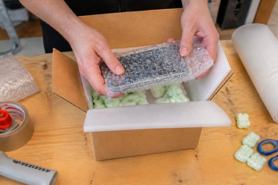 Hands carefully wrapping a smartphone in protective bubble wrap and foam padding inside a cardboard shipping box with cushioning materials
