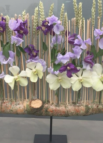 Décoration à poser, socle noir en métal, branche d'arbre à l'horizontal, avec des épis de blé piqué à la verticale, des fleurs d'orchidées écru et violettes sont accrochées aux épis de blé.