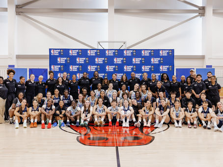 Large mixed basketball group poses in a bright gym before a Basketball Without Borders banner, smiling in team uniforms for Women’s BWB Next Up 2026.