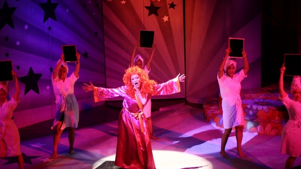 Theatrical scene with a red-haired performer as Zsazsa Zaturnnah in a pink gown, arms outstretched, flanked by dancers holding black tablets under purple-red lights.