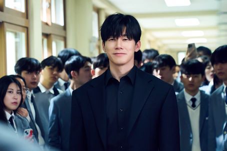 Kim Moo-yul in a black suit stands in a school hallway, calm and confident, surrounded by uniformed students.