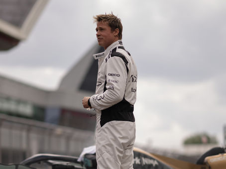 Brad Pitt as Sonny Hayes, a race car driver in a white suit standing beside a car on a cloudy day at a racetrack. Logos visible on suit. Focused expression.