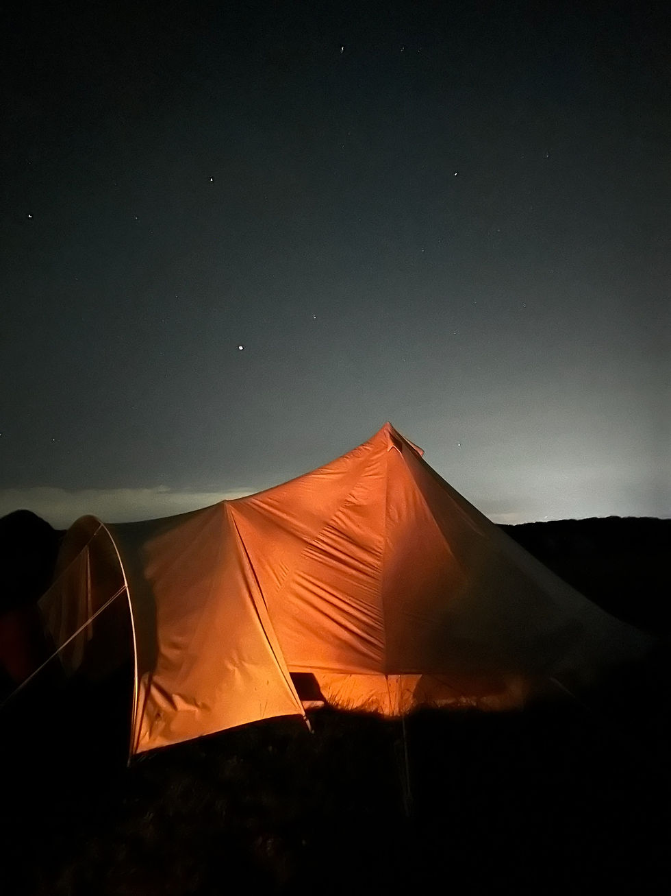 A bell tent at night, lit up by the glow of a campfire with the night sky in the background.