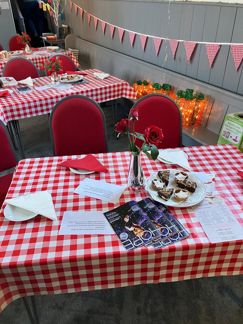 A close-up of a table set with a red and white checkered cloth, featuring mince pies, red roses, and