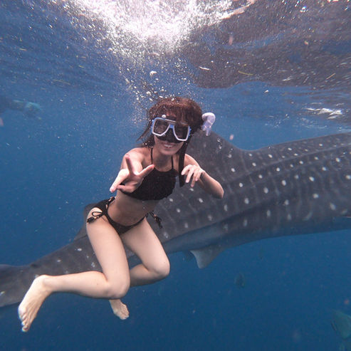 Tourist underwater in Oslob wearing snorkeling gear and posing with a peace sign, with a whale shark behind her.