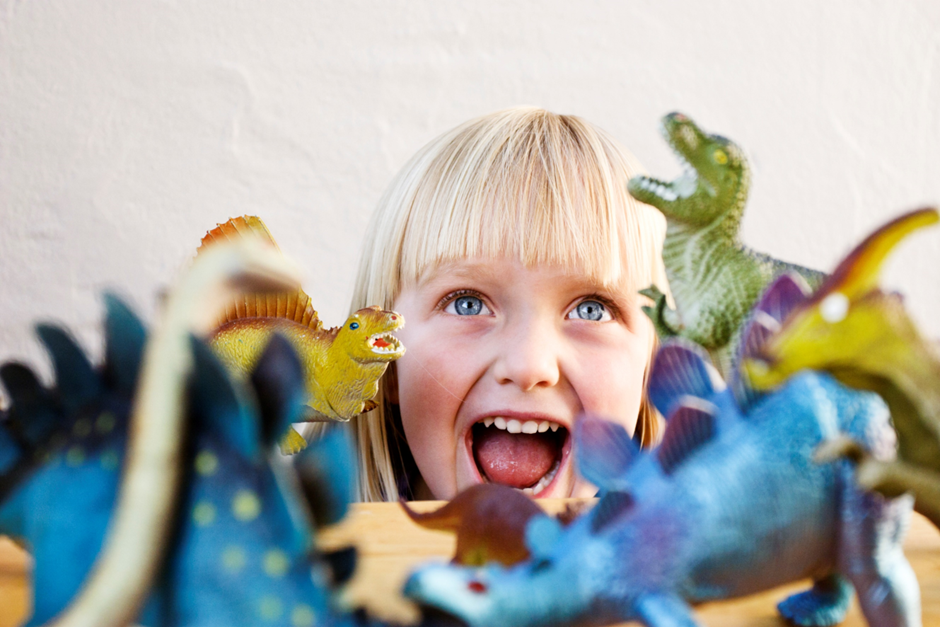 A blonde girl sits behind plastic dinosaurs, roaring at the camera.