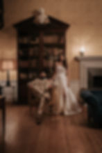 Couple in formal attire, with man seated and woman standing in an elegant room within Botleys Mansion with a vintage bookshelf, warm lighting, and ornate wallpaper.