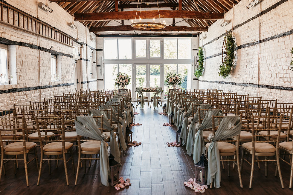 Wooden chairs line a rustic wedding aisle with green drapes and pink petals. Floral arrangements and greenery decorate a bright, airy room at Lillibrooke Manor.