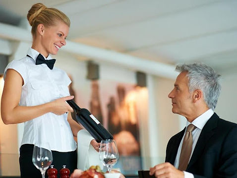 Smiling waitress presenting a bottle of wine to a seated male guest in a formal dining setting, highlighting professional hos