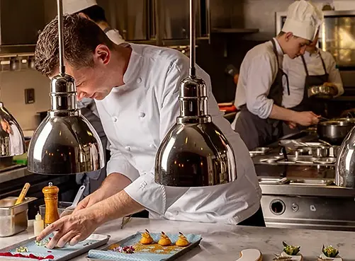 Chef in a professional kitchen carefully plating gourmet dishes under heat lamps, with another chef working in the background