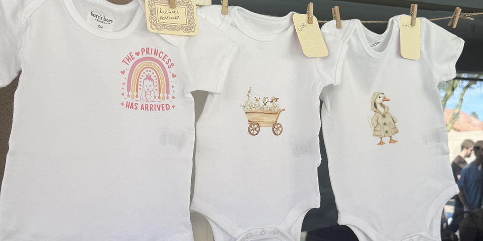 Three white baby onesies hang on a clothesline with tags; prints show a princess, cart, and duck in a soft outdoor market scene.