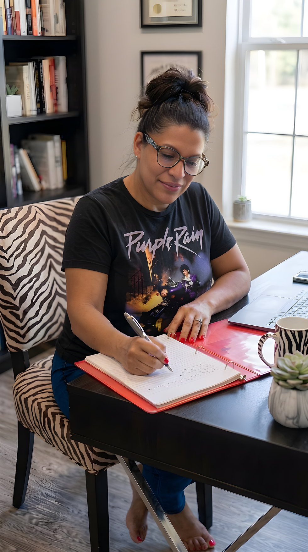 Woman in glasses writes in a notebook at a home desk beside a laptop; her Purple Rain T-shirt, zebra chair, and window-lit room show.