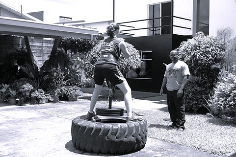 Addo the Coach observing a client doing and exercise. The client is doing belt squats while standing on a tractor tire. Weights can be seen attached to the belt.
