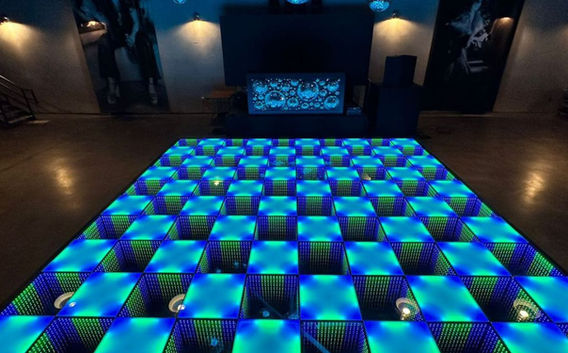 A modern, dimly lit event space showcasing a blue and green LED dance floor with a checkerboard pattern, complemented by hanging mirrored disco balls and a large screen in the background, creating a high-energy atmosphere.