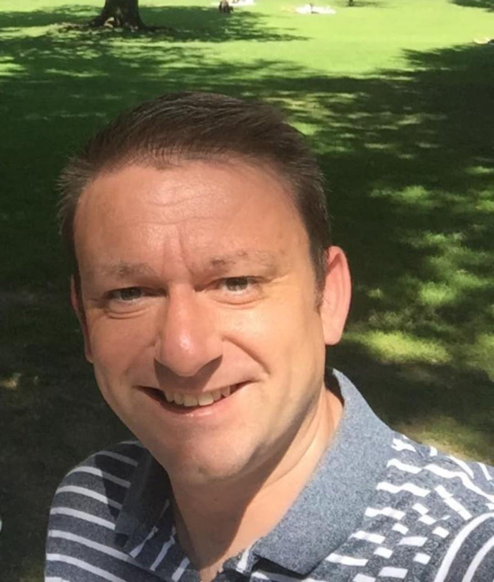 Man smiling in striped polo in sunlit park, lush green grass, trees casting shadows. Bright and cheerful mood.