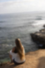 Eine blonde Frau, die auf einer Klippe sitzt, mit dem Rücken zur Kamera, einer weißen Bluse trägt und aufs Meer schaut. 