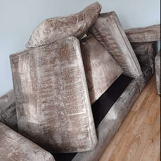 A disassembled brown velvet couch, cushions stacked haphazardly, rests on a wooden floor