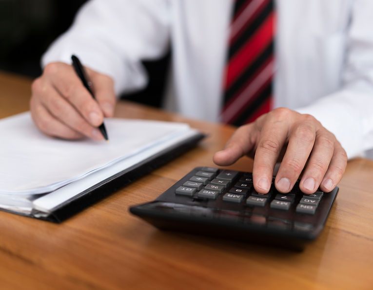 A person in a white shirt and red-striped tie uses a calculator and writes on paper at a wooden desk
