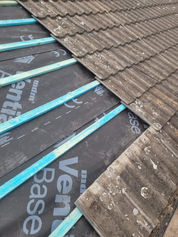 Close-up of a roof under renovation, showing exposed wooden beams and new underlayment material with old gray tiles partially removed.