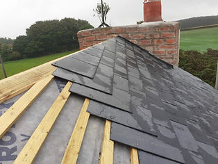 Slate tiles partially installed on a wooden roof near a brick chimney