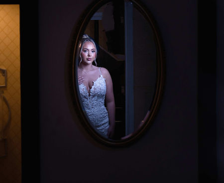 A bride in a white lace gown, with an elegant updo, is reflected in an oval mirror against a dark background