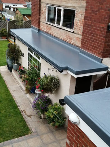 Flat grey roofing on a red-brick house extension, with potted plants and flowers along the patio.