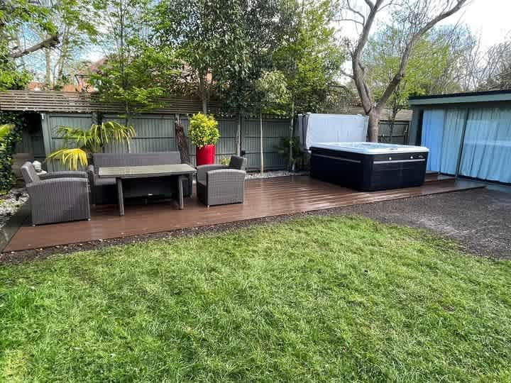 Black hot tub and grey rattan furniture on a wooden garden deck overlooking a green lawn and trees