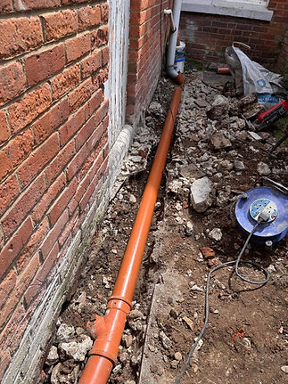 A long orange drainage pipe lies in a trench beside a red brick wall