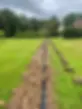 A freshly dug trench cuts across a lush green lawn under a cloudy sky