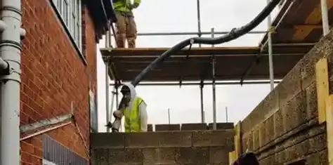 Workers in high-vis vests using a concrete pump hose to fill grey concrete block walls on a construction site