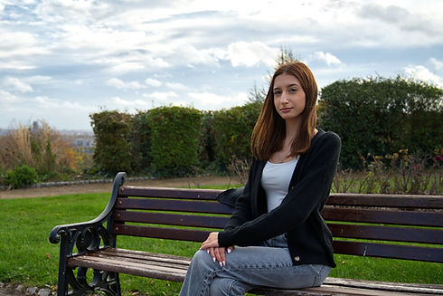 Woman with long hair in a dark jacket sitting on a wooden park bench outdoors with green trees and a cloudy sky