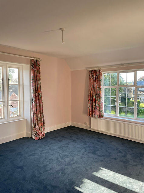 An empty room with pale pink walls and dark carpet features two large windows with floral curtains