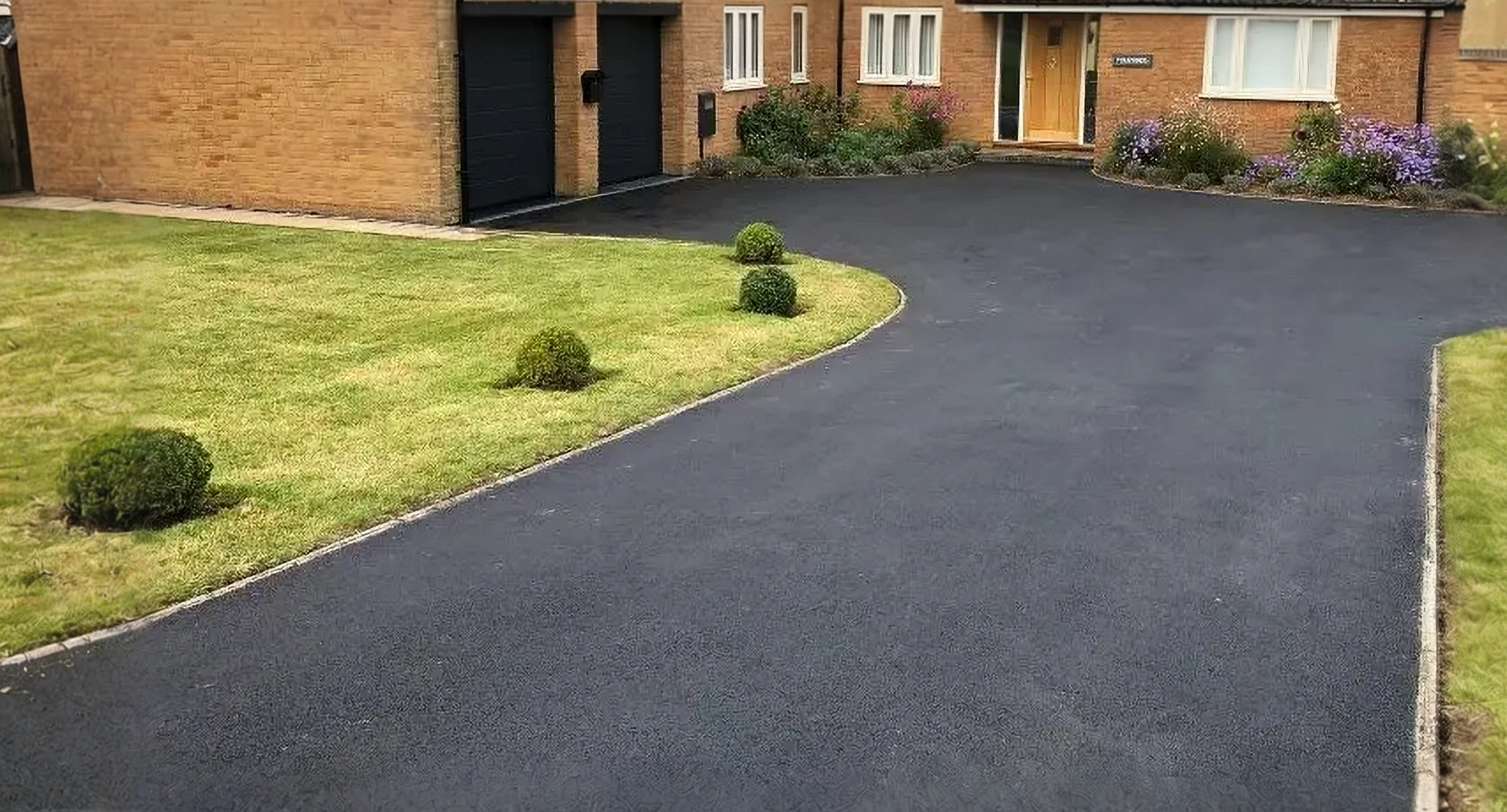 Curving black tarmac residential driveway with block paving borders next to a green lawn
